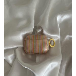 May include: A striped AirPod case in shades of pink, yellow, and blue with a yellow ring clasp. The case is set against a white, silky background.
