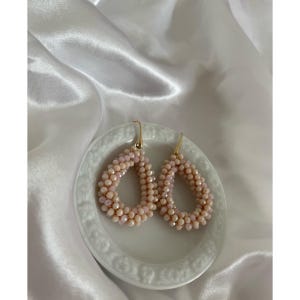 May include: A pair of teardrop-shaped earrings. The earrings are made of small, light pink beads and have gold-colored hooks. They are displayed on a small, white, decorative plate against a white, satin background.