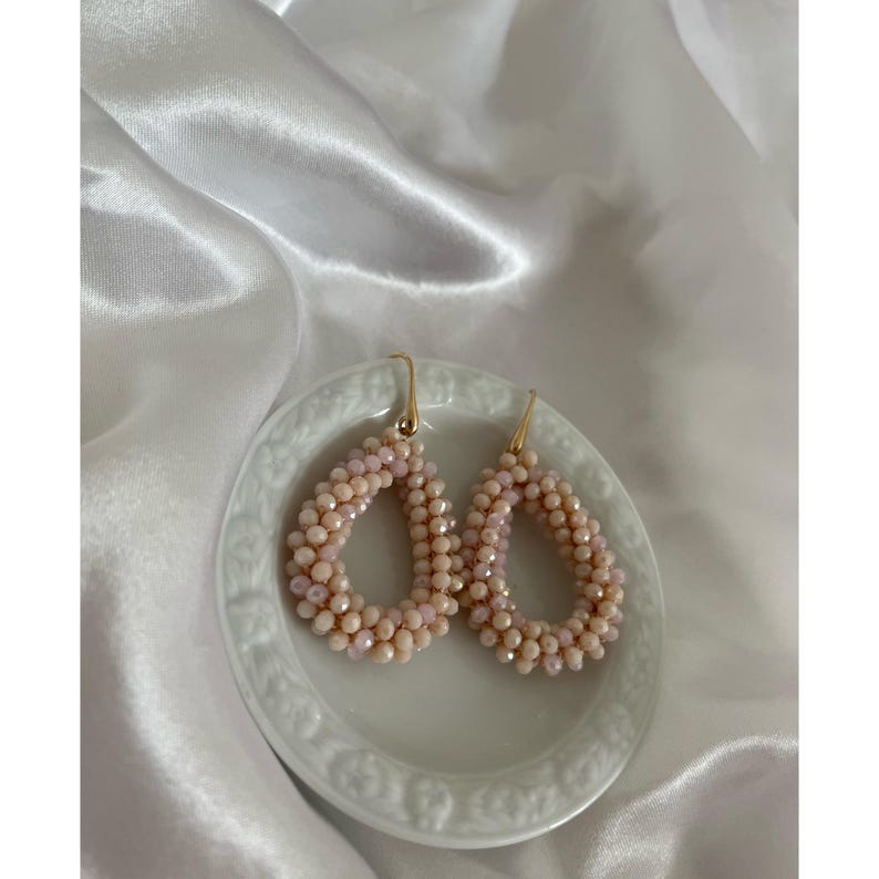 May include: A pair of teardrop-shaped earrings. The earrings are made of small, light pink beads and have gold-colored hooks. They are displayed on a small, white, decorative plate against a white, satin background.