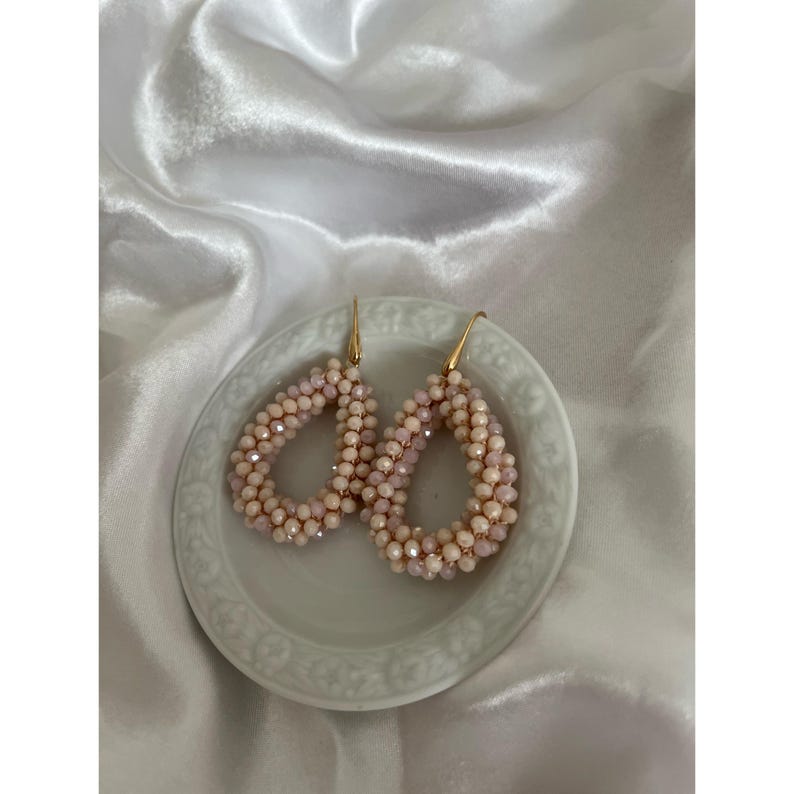 May include: A pair of teardrop-shaped earrings with a beaded design. The earrings are light pink and cream-colored, with gold-colored hooks. They are displayed on a small, white, decorative plate.