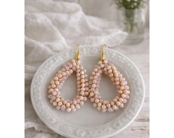 May include: A pair of teardrop-shaped earrings with a beaded design in shades of peach and cream. The earrings are displayed on a white plate with a gold-colored hook. The background includes a white cloth and a vase of flowers.