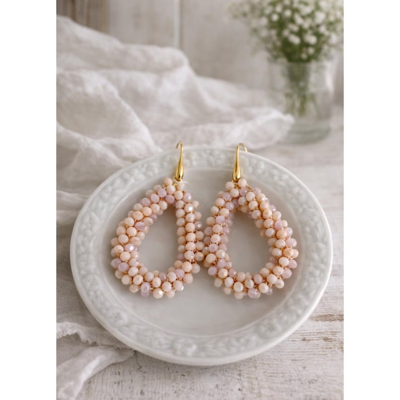 May include: A pair of teardrop-shaped earrings with a beaded design in shades of peach and cream. The earrings are displayed on a white plate with a gold-colored hook. The background includes a white cloth and a vase of flowers.