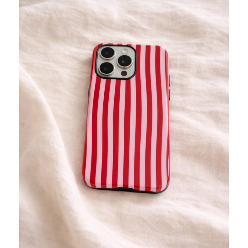 Op de afbeelding: Een rood-roze gestreepte telefoonhoes. De hoes heeft een zwarte rand en bedekt de achterkant en zijkanten van de telefoon. De hoes heeft een verticaal streepjespatroon met afwisselend rode en roze strepen.