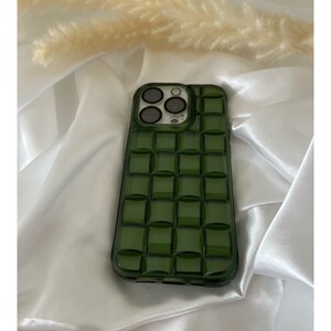 May include: A green phone case with a textured, checkered design. The case has a raised camera bump with three circular cutouts.