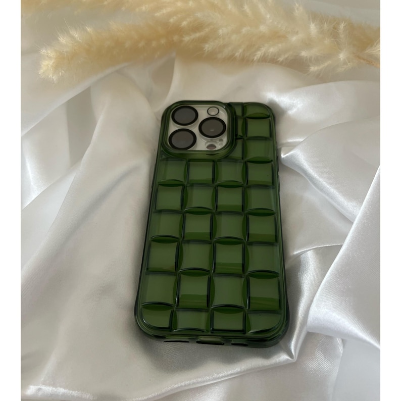 May include: A green phone case with a textured, checkered design. The case has a raised camera bump with three circular cutouts.