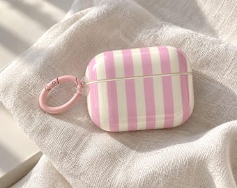 May include: A rectangular protective case for wireless earbuds featuring a vertical striped pattern in light pink and cream. The case includes a matching light pink circular carabiner clip attached to the side for secure carrying and easy attachment.