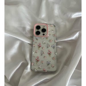 Pode incluir: Capa de telefone transparente com um padr&atilde;o floral de flores rosa, brancas e roxas. A capa tem uma borda rosa ao redor da lente da c&acirc;mera. A capa &eacute; projetada para um smartphone.