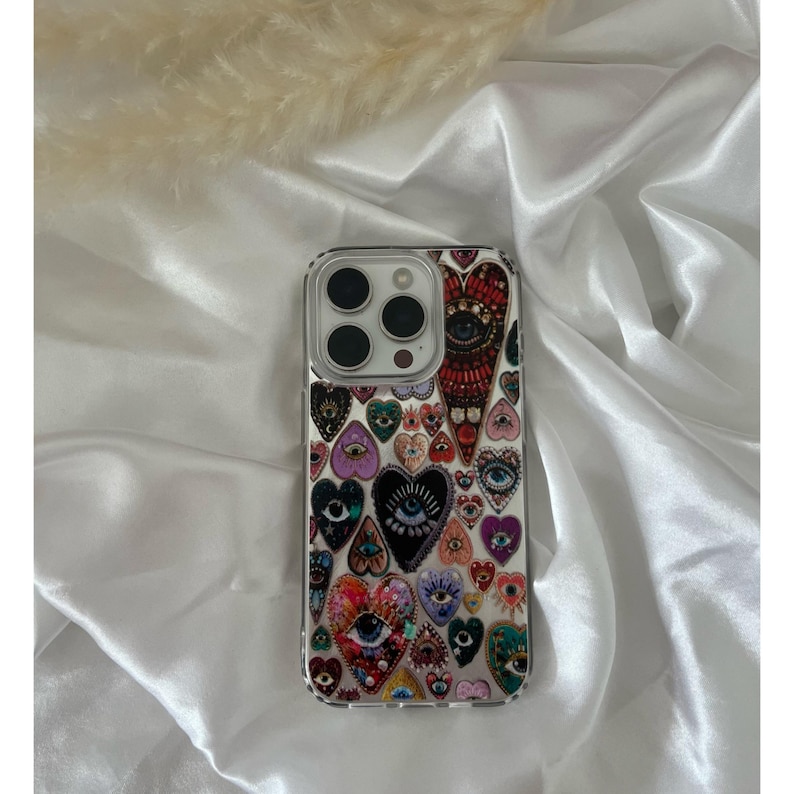 May include: Clear phone case with a colorful heart and eye pattern. The design features various heart shapes with eye illustrations in shades of red, purple, blue, and green. The case has a clear border and a camera cutout.