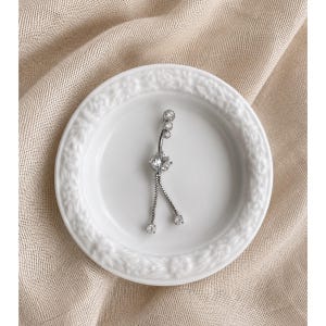 Könnte beinhalten: Ein silberfarbener Bauchnabelpiercing mit klaren, funkelnden Edelsteinen. Der gebogene Stab hat oben eine Ansammlung kleiner Steine, in der Mitte einen größeren Stein und unten hängende Steine. Der Schmuck liegt auf einem weißen Teller.