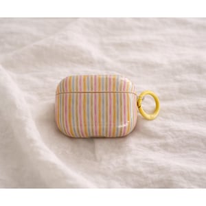 Könnte beinhalten: Ein gestreiftes AirPods-Case in Pastellfarben Gelb, Rosa und Blau. Das Case hat einen gelben Ring zur Befestigung und liegt vor einem weißen, strukturierten Hintergrund. Das Case dient zum Schutz und Transport von kabellosen Ohrhörern.