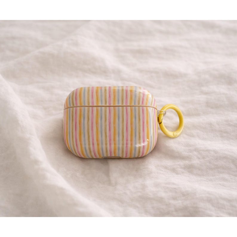 May include: A striped AirPods case in pastel colors of yellow, pink, and blue. The case has a yellow ring for attachment and is set against a white, textured background. The case is designed to protect and carry wireless earbuds.