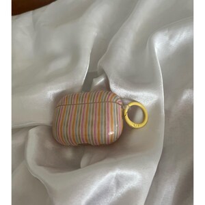 May include: A striped AirPods case in shades of pink, yellow, and orange with a yellow ring clasp. The case has a glossy finish and is set against a white, silky background.