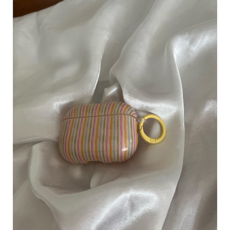 May include: A striped AirPods case in shades of pink, yellow, and orange with a yellow ring clasp. The case has a glossy finish and is set against a white, silky background.