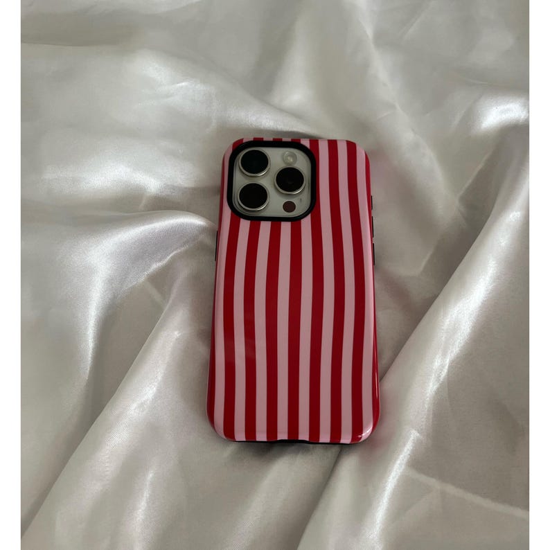 Op de afbeelding: Een rood-roze gestreepte telefoonhoes. De hoes heeft een glanzende afwerking en een zwarte rand rond de cameralenzen. De achtergrond is een witte, zijdeachtige stof.