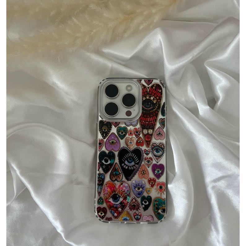 May include: Clear phone case with a colorful heart and eye design. The case features a variety of heart shapes with eye illustrations in various colors, including red, purple, and blue. The case is designed to fit a smartphone.