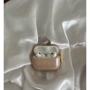 May include: White wireless earbuds inside a striped case with pink, yellow, and gray vertical stripes. The case is open, revealing the earbuds. The case has a yellow clasp and is set against a white, silky background.