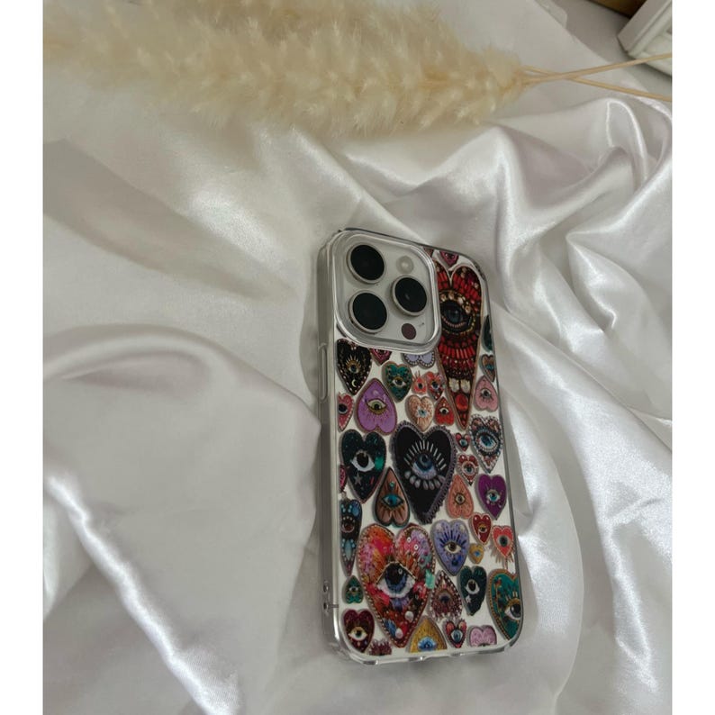 May include: Clear phone case with a colorful heart and eye design. The case features a variety of heart shapes with intricate eye patterns in shades of red, purple, blue, and green. The case is designed to fit a smartphone.