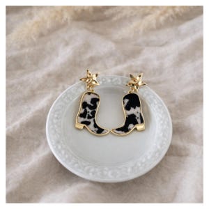 Könnte beinhalten: Ein Paar goldfarbene Ohrringe mit einem Cowboy-Stiefel-Design mit einem schwarz-weißen Kuhfellmuster. Jeder Ohrring hat eine sternförmige Spitze. Die Ohrringe werden auf einem weißen, dekorativen Teller präsentiert.