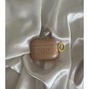 May include: A striped AirPod case with a yellow clasp. The case features vertical stripes in shades of pink, yellow, and gray. The case is on a white satin surface.