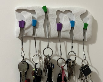 May include: A white 3D printed key holder with six hooks. Each hook has a colourful plastic clip and a metal chain. The hooks are holding a variety of keys, including car keys, house keys, and a carabiner.