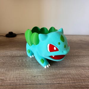 May include: A turquoise Bulbasaur planter with green detailing, including the leaves on its back and the spots on its body. The planter has red eyes and a red mouth. The planter is made of a 3D printed material.