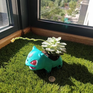 Può includere: Un vaso da fiori verde e blu a forma di Pokemon con una pianta grassa che cresce al suo interno. Il vaso ha la forma di un personaggio Pokemon con occhi rossi e una bocca bianca.