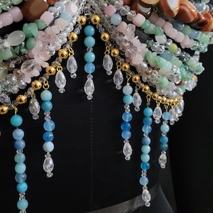 May include: A close-up of a beaded garment with a variety of colorful gemstones. The piece features strands of blue, pink, green, and orange beads, accented with gold-colored metal and clear crystal teardrops. The garment is displayed on a black mannequin.