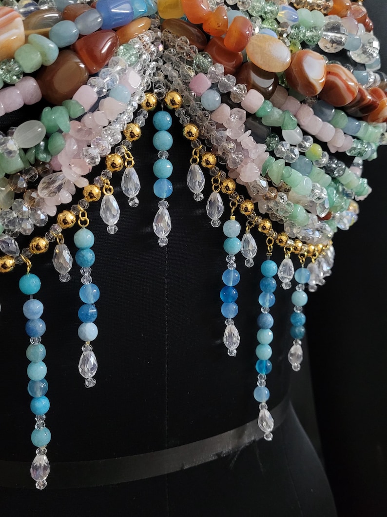 May include: A close-up of a beaded garment with a variety of colorful gemstones. The piece features strands of blue, pink, green, and orange beads, accented with gold-colored metal and clear crystal teardrops. The garment is displayed on a black mannequin.