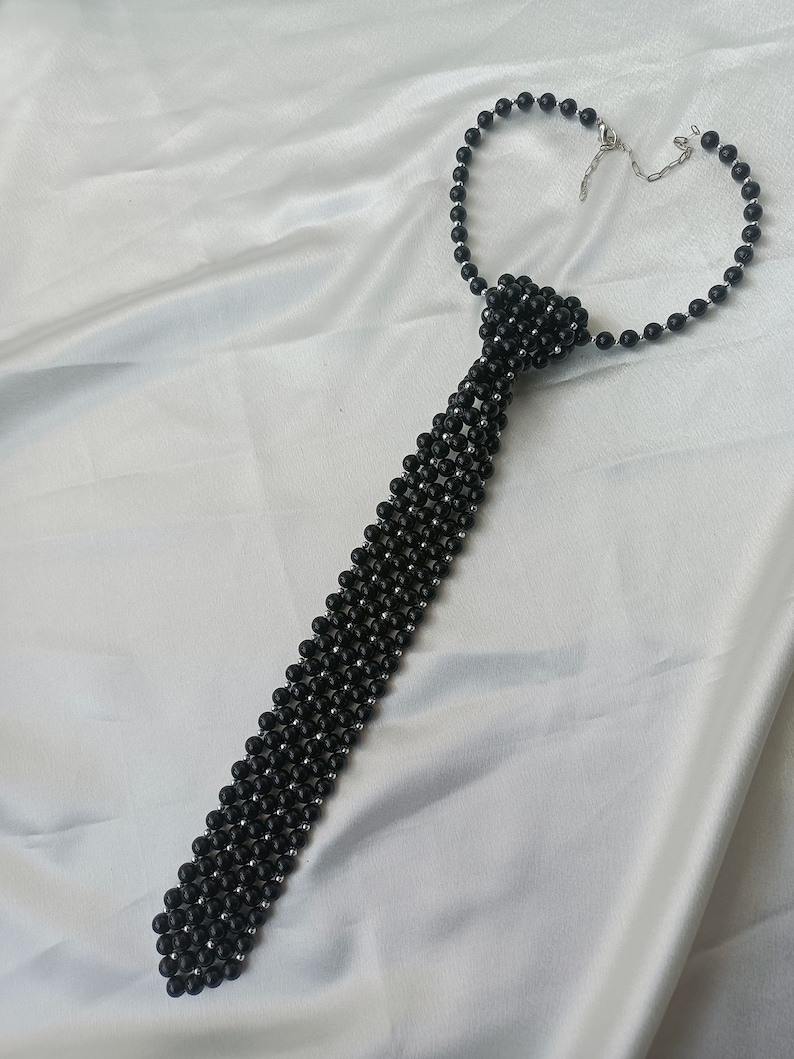 Pu&ograve; includere: Una collana a cravatta con perline nere e chiusura argentata. Le perline sono disposte in un motivo che ricorda una cravatta.