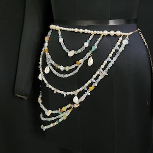May include: A multi-strand beaded body chain with white, clear, and light green beads, seashells, and gold accents. The chain is worn on a black mannequin.