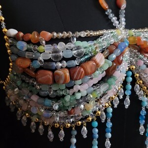 May include: A multi-strand beaded top featuring a variety of colorful gemstones and clear beads. The design incorporates shades of orange, blue, green, and pink, with gold-colored accents and dangling crystal-like embellishments. The top is set against a black background.