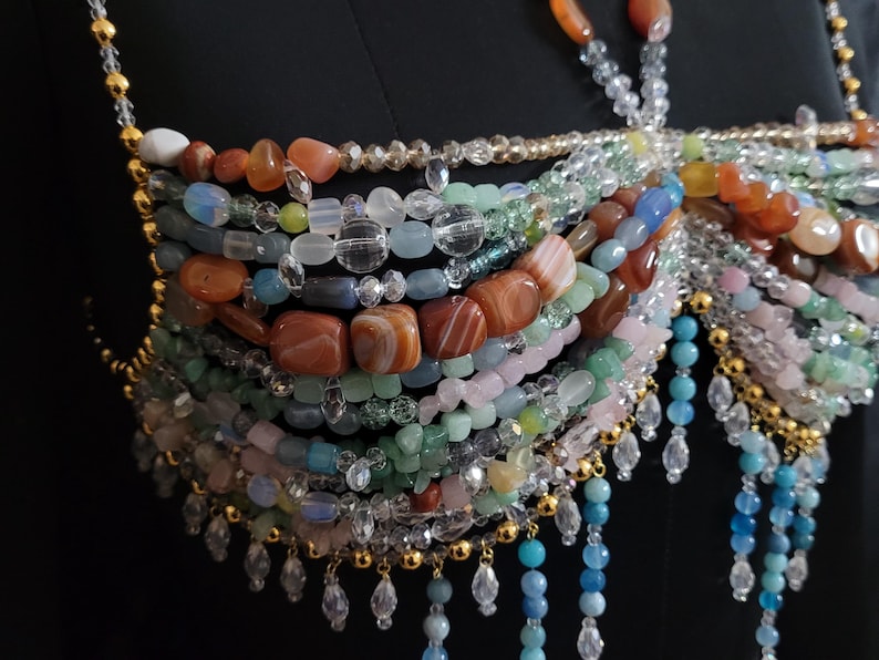 May include: A multi-strand beaded top featuring a variety of colorful gemstones and clear beads. The design incorporates shades of orange, blue, green, and pink, with gold-colored accents and dangling crystal-like embellishments. The top is set against a black background.