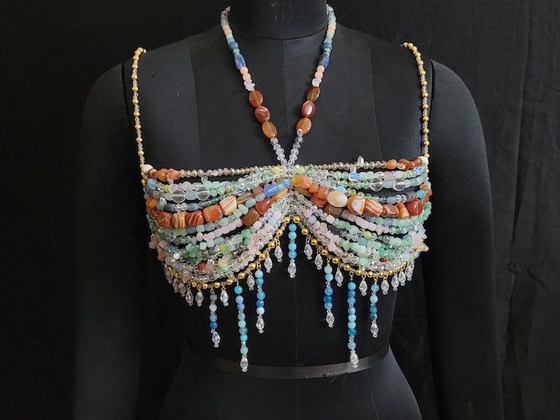 May include: A beaded top in various colors, including orange, green, pink, and blue, is displayed on a black mannequin. The top features multiple strands of beads, with a halter-style neckline and dangling bead accents.