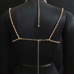 May include: A gold-toned body chain with a beaded necklace and shoulder straps. The straps are adorned with small gold beads and connect to a horizontal chain across the back. A longer chain hangs down the back, ending with a small crystal.