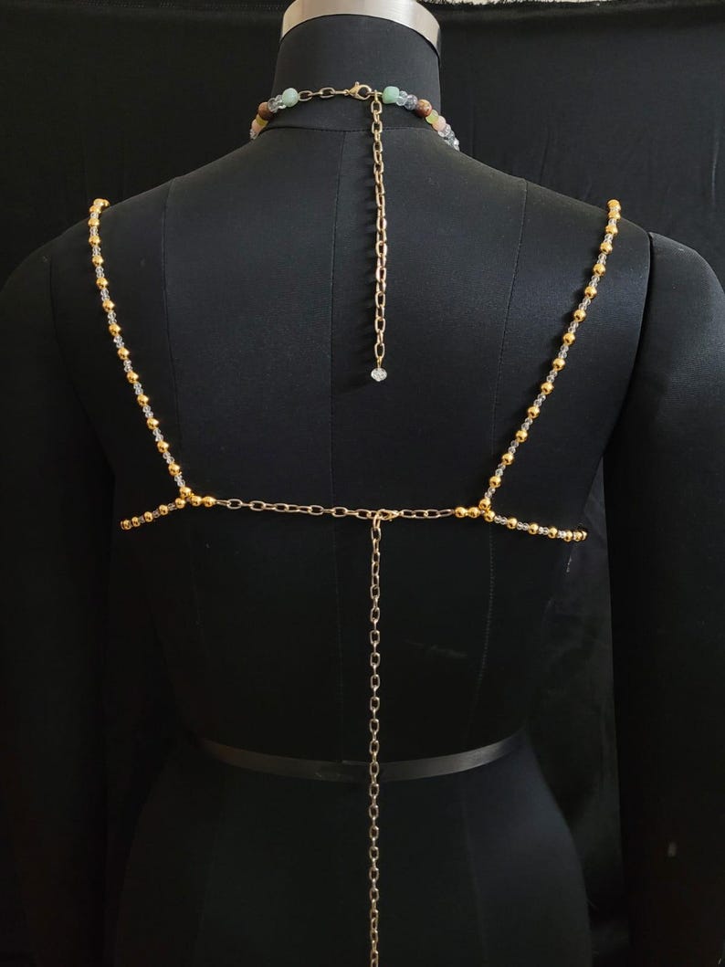 May include: A gold-toned body chain with a beaded necklace and shoulder straps. The straps are adorned with small gold beads and connect to a horizontal chain across the back. A longer chain hangs down the back, ending with a small crystal.