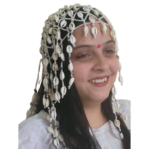 May include: A white shell headdress with dangling shell accents. The headpiece is adorned with small white shells and beads, creating a delicate and ornate design. The shells are arranged in a pattern across the head and cascading down the sides.
