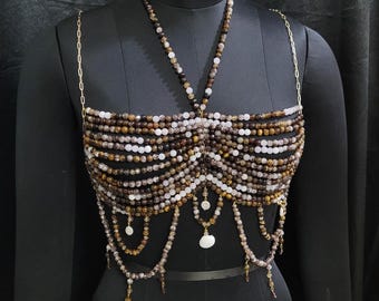Könnte beinhalten: Ein Perlenoberteil aus braunen, weißen und goldenen Perlen. Das Oberteil hat einen Halterneck-Ausschnitt und ein mehrschichtiges Design. Die Perlen sind auf einer goldenen Kette aufgefädelt.