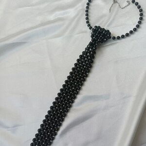 Pu&ograve; includere: Una collana a cravatta con perline nere e chiusura argentata. Le perline sono disposte in un motivo che ricorda una cravatta.