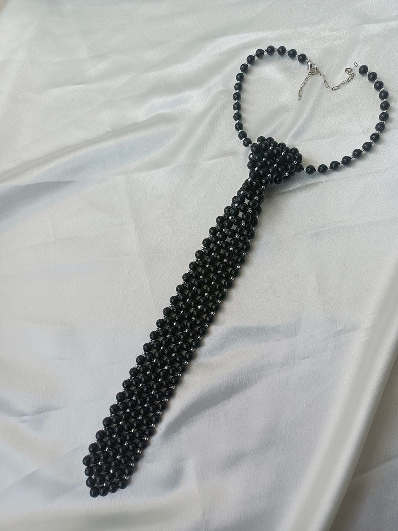 Pu&ograve; includere: Una collana a cravatta con perline nere e chiusura argentata. Le perline sono disposte in un motivo che ricorda una cravatta.