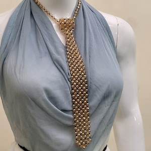 Peut inclure: Un collier cravate en perles dorées et noires. La cravate est faite de petites perles rondes et est présentée sur un mannequin. Le fond est un tissu bleu clair.