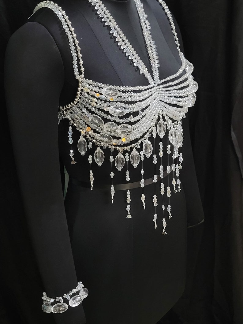 May include: A clear crystal beaded top with a delicate, intricate design. The top features a variety of crystal shapes and sizes, including small round beads, larger teardrop-shaped beads, and long, dangling strands. The top is designed to be worn as a statement piece and would be perfect for a special occasion.
