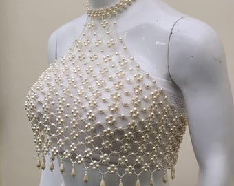 May include: A cream pearl halter top and choker necklace. The top has a net-like design with a floral pattern. The bottom edge features dangling pearl accents. The top is displayed on a white mannequin.