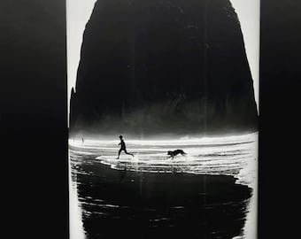 Pode incluir: Fotografia em preto e branco de uma pessoa e um cão correndo em uma praia com uma grande formação rochosa ao fundo. A fotografia é impressa em um copo branco com a assinatura do fotógrafo na parte inferior.