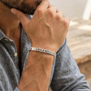 Puede incluir: Una pulsera de plata con un diseño de cadena de eslabones. La pulsera tiene una placa rectangular con texto grabado. La persona de la imagen lleva una camisa gris abotonada.