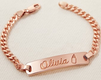May include: A rose gold baby bracelet with a curb chain and a personalized nameplate engraved with the name "Olivia" and a baby bottle icon.