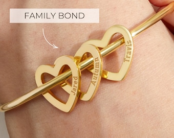 May include: Gold bangle bracelet with three interlocking heart charms. Each heart charm has a name engraved on it: Jared, Autumn, and Travis. The bracelet is on a person's wrist.