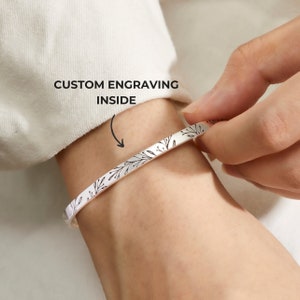 Op de afbeelding: Een zilveren armband met een bloemenmotief gegraveerd aan de buitenkant. De tekst "CUSTOM ENGRAVING INSIDE" is zichtbaar boven de armband.