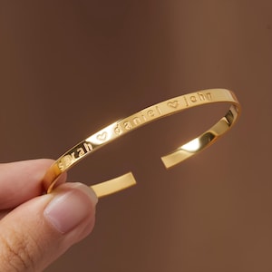 Puede incluir: Una pulsera de brazalete abierta de color dorado. La pulsera está grabada con los nombres "sarah", "daniel" y "john", separados por símbolos de corazón. La pulsera tiene un acabado liso y pulido y está sujeta por una mano.