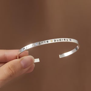 May include: A silver-colored open cuff bracelet with the names "maria", "anne", and "audrey" engraved on it. The bracelet has small, colorful gemstones between the names. The bracelet is held by a hand.