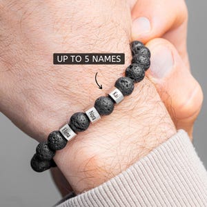 Op de afbeelding: Zwarte lavasteen kralenarmband met zilveren naamkralen. De armband heeft de namen "Elena", "Nora" en "Kai" gegraveerd op de zilveren kralen. De tekst "UP TO 5 NAMES" staat erboven.